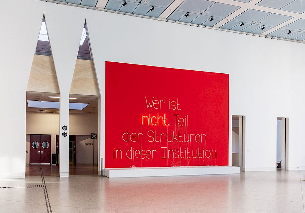 Wandinstallation von Lerato Shadi. Auf einer quadratischen roten Fl&auml;che steht in wei&szlig;er Schrift: &bdquo;Wer ist nicht Teil der Strukturen der Institution&ldquo;. Das Wort &bdquo;nicht&ldquo; blinkt auff&auml;llig in Orange.