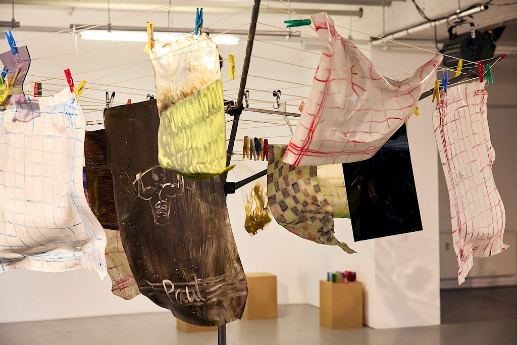 Kunstinstallation von Anna Sch&ouml;l&szlig; mit dem Titel Heat it up (2025). Zu sehen ist eine W&auml;schespinne im Ausstellungsraum, an der mit bunten W&auml;scheklammern bemalte und bearbeitete Kunststoff- oder Textilbahnen h&auml;ngen. Die Bahnen zeigen Gittermuster, abstrakte Farbverl&auml;ufe und Motive, darunter eine gezeichnete Figur mit der Aufschrift &bdquo;Paul&ldquo;. Die Objekte h&auml;ngen in verschiedenen H&ouml;hen und Formen an gespannten Leinen. Im Hintergrund sind Podeste mit farbigen Objekten sowie eine wei&szlig;e Wand und Decke mit sichtbarer Technik zu erkennen. Die Szene ist hell beleuchtet.