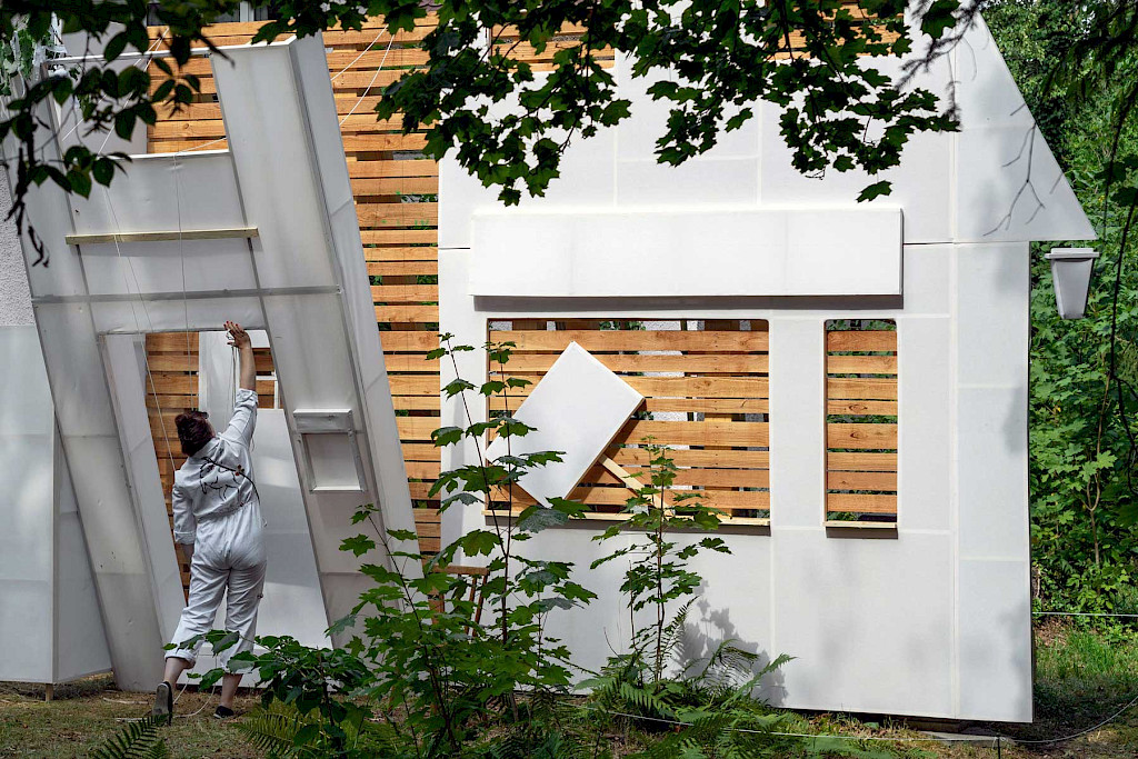 Foto einer Arbeit von Gabi Blum. Eine weiblich gelesene Person stellt eine Fassade von einem Haus auf. Die Fassade ist wei&szlig;.