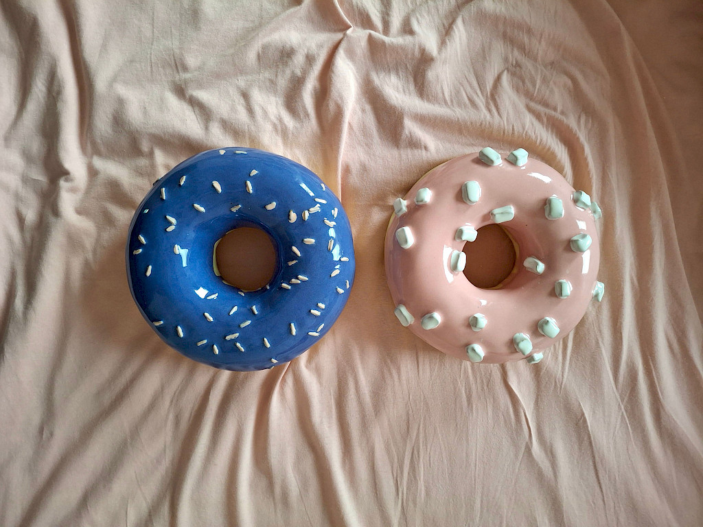 Fotografie von Clara Umbach. Auf einer beigen Bettdecke liegen zwei Donuts aus Keramik. Ein Donut hat eine blaue Glasur mit wei&szlig;en Streuseln. Der andere Donut hat eine rosafarbene Glasur mit wei&szlig;en kleinen Marshmallows.